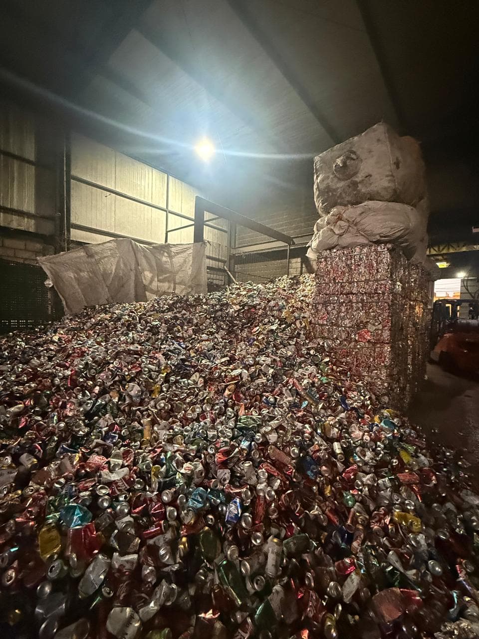 Gran pila de latas de aluminio trituradas en un almacén con fardos de reciclaje apilados.