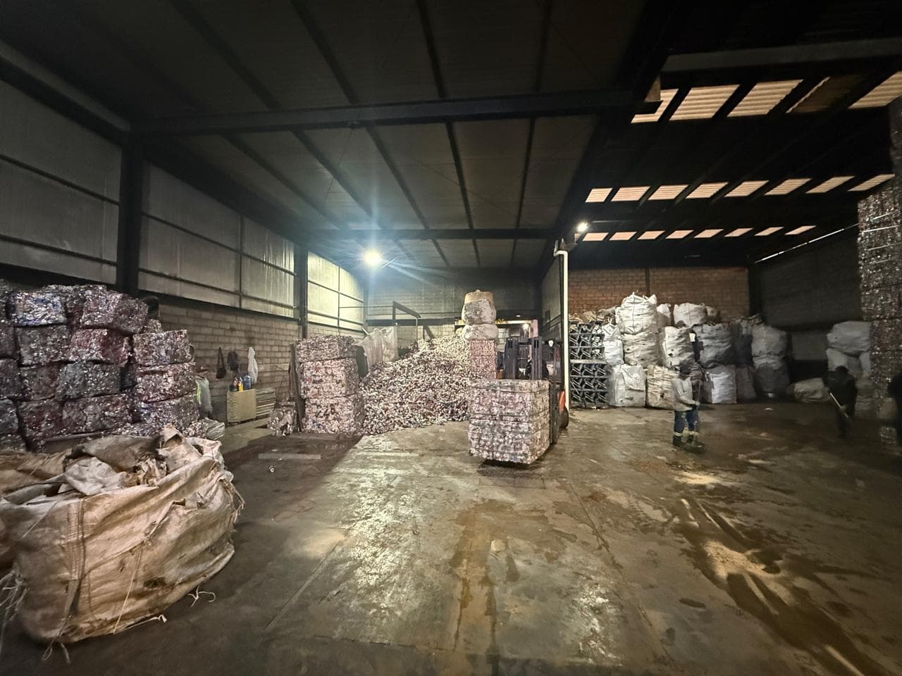 Interior de planta de reciclaje con montacargas moviendo pacas de metal entre pilas de materiales.