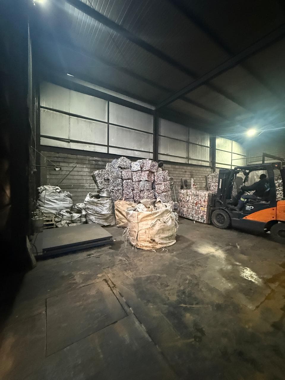 Montacargas naranja moviendo fardos compactados en un almacén de reciclaje con grandes sacos blancos.
