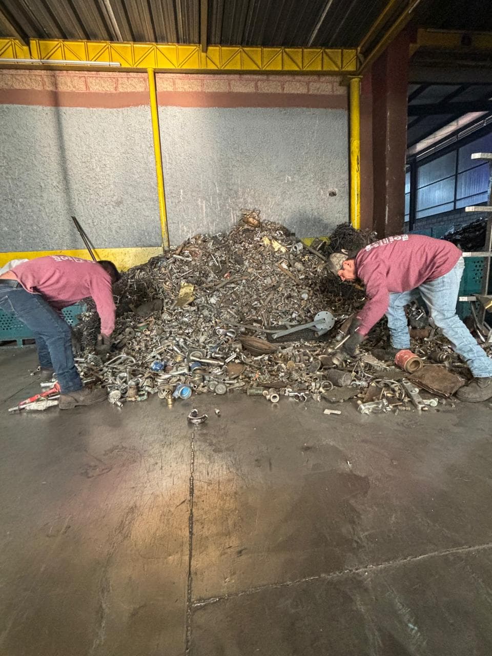 Dos trabajadores clasifican manualmente una gran pila de chatarra metálica en un almacén industrial.