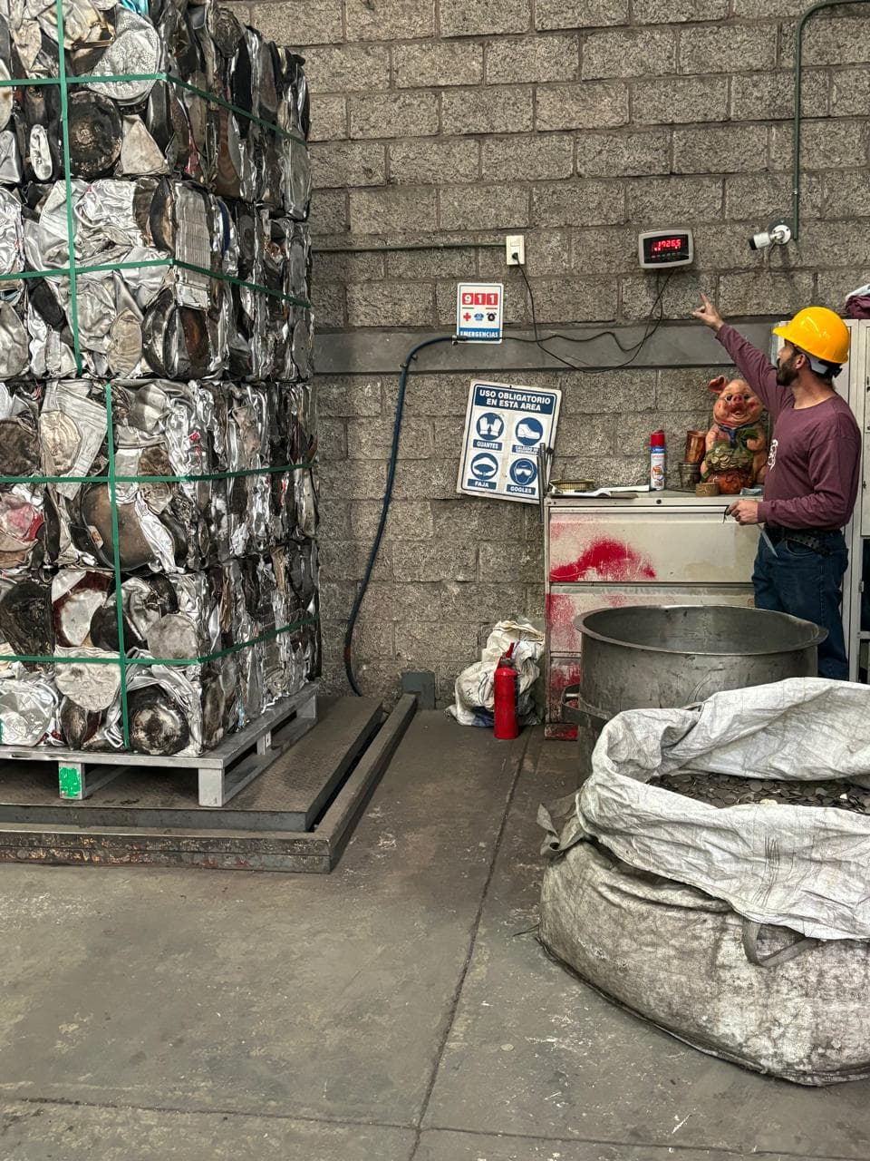 Operario con casco amarillo señala la báscula digital pesando una paca de metal reciclado.